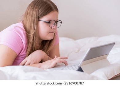 Positive overweight teenage girl looking in tablet using touch-pad on wireless keyboard teenager learning school subjects online from home lying on bed in room - Powered by Shutterstock