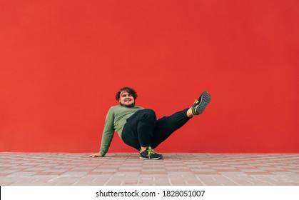 Positive Overweight Young Man Dancing Hip Hop On The Street On A Red Wall Background. Tostun Shows A Dance Ball Performance On A Red Background.