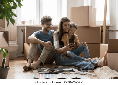 Positive parents and happy kids taking moving selfie on smartphone, sitting at stacked cardboard boxes, renovation brochures with wall color samples, holding mobile phone, smiling - Powered by Shutterstock