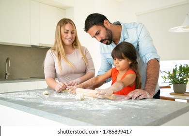 Positive Parents Watching Daughter Rolling Dough On Kitchen Desk With Flour Messy. Young Couple And Their Girl Baking Buns Or Pies Together. Family Cooking Concept