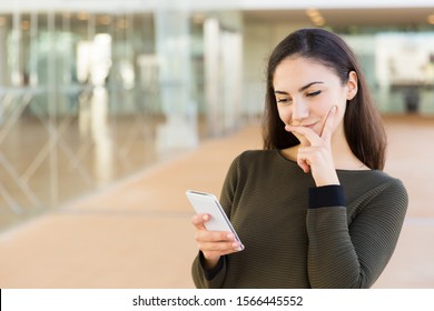 Positive Pensive Latin Woman With Cellphone Touching Face And Reading Message. Young Woman With Mobile Phone Posing Indoors With Glass Wall Interior In Background. Mobile Communication Concept