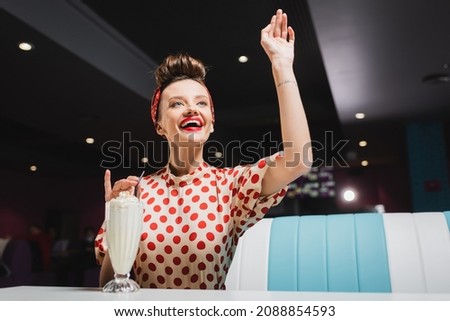 positive pin up woman in red polka dot blouse gesturing near milkshake on table