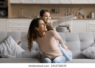 Positive playful pretty mom piggybacking little tween daughter with open arms. Sweet girl playing flying airplane on mothers back, enjoying activity on home sofa