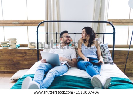 Positive playful woman teasing smiling content boyfriend showing tongue during spending free time together in home bed with modern devices