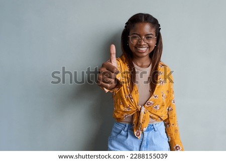 Positive portrait of young African American woman gesturing thumb up, standing against a gray wall, looking at camera smiling. Isolated background. Copy space image.