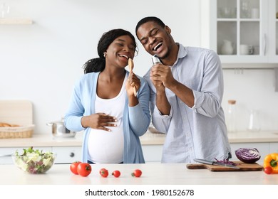 Positive Pregnant Black Couple Singing While Cooking At Home, Happy African American Expecting Lady Making Dinner With Her Husband, Singing Songs And Dancing, Kitchen Interior. Family, Love Concept