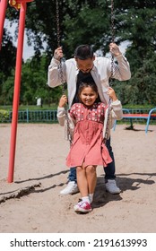 Positive Preteen Girl Sitting On Swing Near Asian Dad In Park