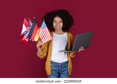 Positive Pretty Black Child Girl With Bushy Hair Holding Computer And Bunch Of Flags Of Various Countries, Burgundy Studio Background. Foreign Language Online School Concept