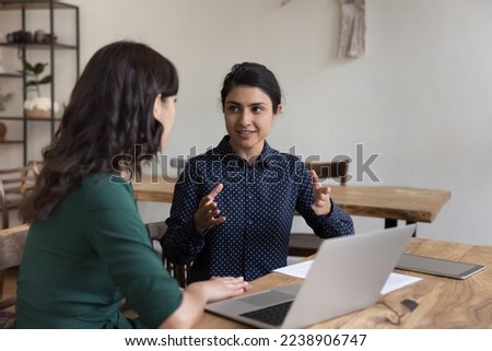 Positive pretty Indian mentor woman training intern, new employee, talking to coworker at office workplace, speaking with hands gestures, explaining task, telling about business project
