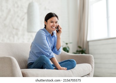 Positive pretty millennial korean lady sitting on sofa at home, talking on mobile phone and smiling, enjoying her unlimited cell phone plan, looking at copy space for advertisement