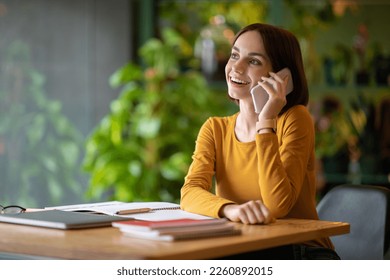 Positive pretty young businesswoman working at coffee shop, lady sitting at table alone, taking notes, making plan, having phone conversation, looking at copy space and smiling - Powered by Shutterstock