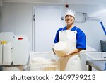 Positive professional female factory worker in uniform showing whole white cheese while standing at work looking at camera