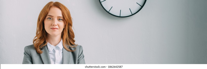 Positive Redhead Businesswoman Looking At Camera Near Wall Clock On Grey, Banner