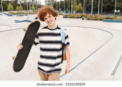 Positive redhead caucasian curly teen school boy guy teenager skater sportsman in casual clothes with bag holding skateboard riding on ramp in skate park after lessons. Summer holiday concept - Powered by Shutterstock