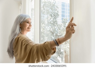 Positive relaxed elder Hispanic homeowner, hotel guest lady parting white transparent curtains, veils at large window, looking at summer city view, enjoying morning sunlight, smiling - Powered by Shutterstock