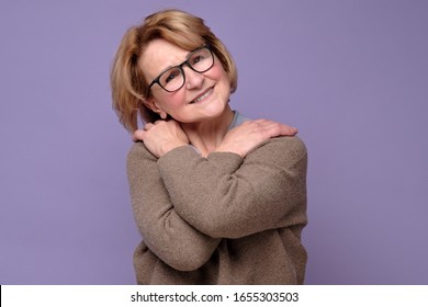 Positive relaxed senior caucasian woman in glasses smiling, keeping arms around herself. Positive facial human emotion.