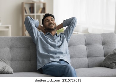 Positive relaxed young Indian man sitting on home sofa, stretching body, leaning on soft back, touching neck with closed eyes, enjoying comfort, coziness, breathing fresh air - Powered by Shutterstock