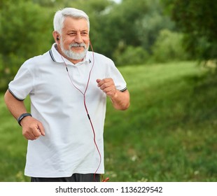 Positive running old man in green city park. Smiling, feeling good. Sport lifestyle. Wearing classic white polo shirt with dark blue stripes, sport watch, red headphones. Trees, grass on background.