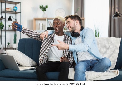 Positive Same Sex Couple In Casual Wear Taking Selfie On Modern Smartphone While Resting Together On Comfy Couch. Happy Loving People Spending Time With Fun At Home.