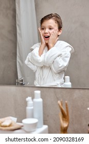 positive school boy applies aftershave like dad in the morning in bath, hygiene at home, young caucasian child looks at mirror reflection surprised. skin care, hygiene, lifestyle concept