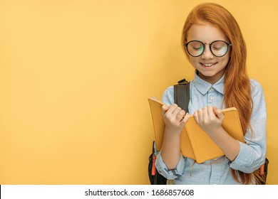 positive school girl with book in hands, red haired child happily reading a book, wearing eye glasses. school, education concept. isolated yellow background