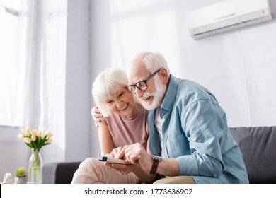 Positive senior couple using remote controller of air conditioner on sofa