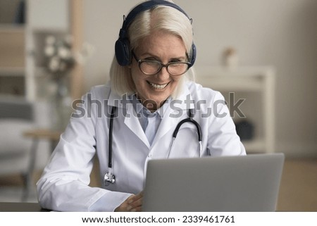 Positive senior doctor woman in wireless headphones speaking on online video call to patient, giving assistance, consultation, phone support. Medical expert giving webinar, lecture on Internet