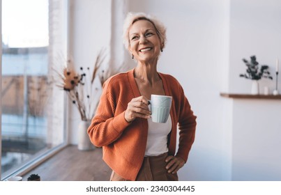 Positive senior female with mug of hot beverage smiling and looking away while standing with hand on hip near window at home - Powered by Shutterstock