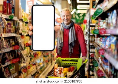 Positive Senior Man Customer Showing Modern Smartphone With White Empty Screen, Walking By Supermarket Aisle With Shopping Trolley Full Of Grocery, Using Mobile Application, Mockup