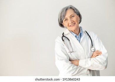 Positive Senior Mature Female Doctor With A Stethoscope In A Medical Coat Posing Isolated Over Gray Background, Standing With Arms Folded, Smiling And Welcoming Healthcare Worker Looking At The Camera