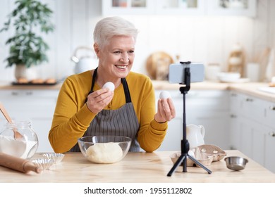 Positive Senior Woman Culinary Vlogger Broadcasting From Home While Baking Cake, Showing Her Followers Eggs, Using Modern Mobile Phone And Tripod, Cozy Kitchen Interior, Copy Space