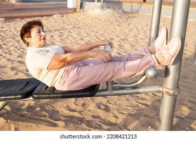 Positive Senior Woman Doing Stretching Outdoors. Pensioner Does Fitness On Beach Using Exercise Equipment. Concept Of Physical Activity In Old Age, Health, Work Out, Healthy Lifestyle. Woman 80+