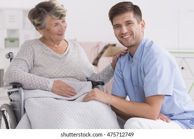 Positive Senior Woman On A Wheelchair And Smiling Male Carer, Light Home Interior