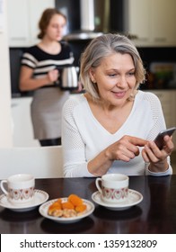 Positive Senior Woman Using Phone While Young Woman Making Tea