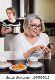 Positive Senior Woman Using Phone While Young Woman Making Tea