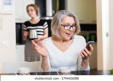 Positive Senior Woman Using Phone While Young Woman Making Tea