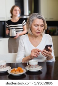 Positive Senior Woman Using Phone While Young Woman Making Tea