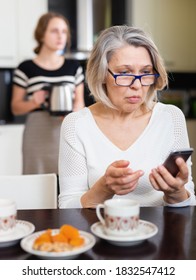 Positive Senior Woman Using Phone While Young Woman Making Tea