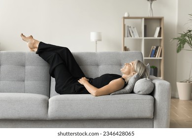 Positive serene mature woman lying on couch with closed eyes and happy smile, placing legs and feet on soft back, enjoying leisure, lazy break at home, breathing fresh cool air - Powered by Shutterstock