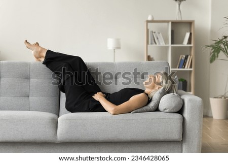Positive serene mature woman lying on couch with closed eyes and happy smile, placing legs and feet on soft back, enjoying leisure, lazy break at home, breathing fresh cool air