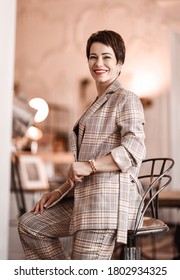 Positive Short Haired Brunette Woman In Elegant Business Plaid Pantsuit With Rolled Up Sleeves Sits On A High Chair In Room Appartement Office Gallery. Business Elegance Concept