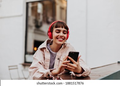 Positive short-haired girl in red headphones smiles outdoors. Brunette woman in beige jacket holding phone and listening to music outside..