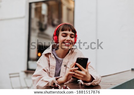 Positive short-haired girl in red headphones smiles outdoors. Brunette woman in beige jacket holding phone and listening to music outside..