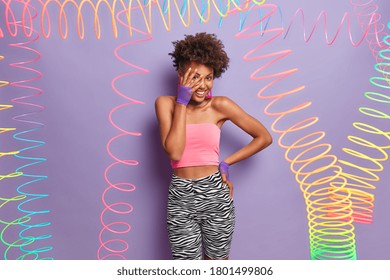 Positive shy curly haired woman makes face palm, wears tank top, leggins and sport gloves, being in good mood, surrounded by colorful slinky, isolated over purple background. Toy plastic rainbow