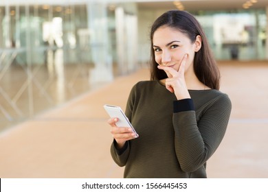 Positive Shy Latin Woman With Cellphone Touching Face And Looking At Camera. Young Woman With Mobile Phone Posing Indoors With Glass Wall Interior In Background. Wireless Technology Concept