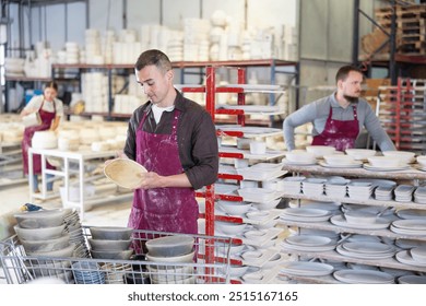 Positive skilled male artisan standing amidst racks with collection of crafted wares in sunlit workshop, meticulously examining slipcast ceramic plates - Powered by Shutterstock