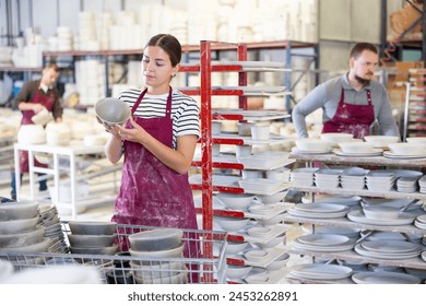 Positive skilled young female artisan standing amidst racks with collection of crafted wares in sunlit workshop, meticulously examining slipcast ceramic plates.. - Powered by Shutterstock