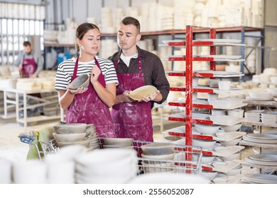 Positive skilled young male and female artisans standing amidst racks with collection of crafted wares in sunlit workshop, meticulously examining and conferring over slipcast ceramic dishes - Powered by Shutterstock