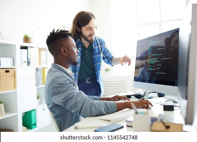 Positive skilled young multiethnic coders in casual clothing discussing computer language: smiling bearded man pointing at computer monitor while explaining web code to African colleague in office