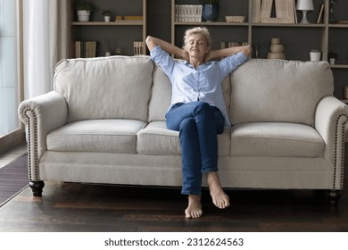 Positive sleepy mature homeowner woman stretching body on soft comfortable couch, relaxing at cozy home, enjoying comfort, relaxation, daydreaming, leisure in modern apartment. Full length shot - Powered by Shutterstock